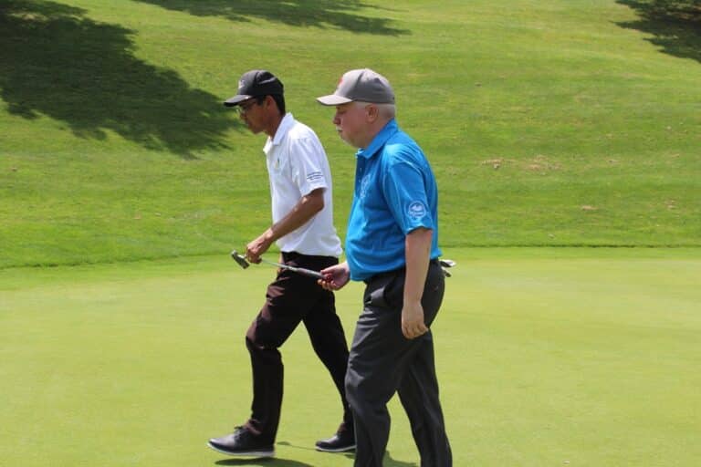 Two golfers are walking on a green approaching the hole. A blind coach in a white shirt, is guiding a B1 golfer using a putter. The putter acts as a stick that helps direct the blind golfer.